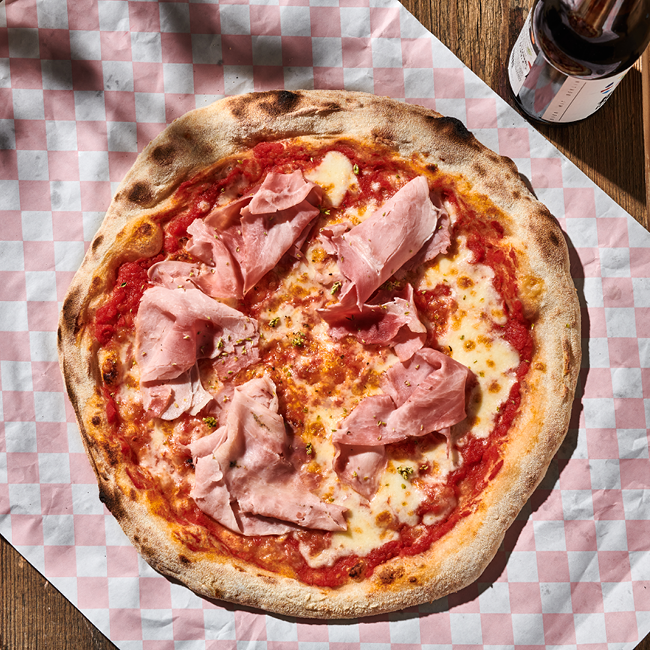 Pizza mit Schinken und Käse auf kariertem Papier, daneben eine Flasche auf einem Holztisch.