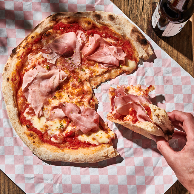 Eine Pizza mit Schinken auf kariertem Papier, eine Hand nimmt ein Stück auf, daneben steht eine Flasche.