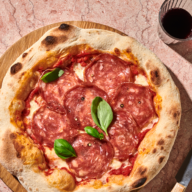 Pizza mit Salami und Basilikumblättern auf einem Holzteller, daneben ein Glas Rotwein auf einem Marmortisch.