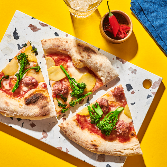 Drei Pizzastücke mit Brokkoli, Wurst und Kartoffelscheiben auf einer Platte, daneben ein Glas Getränk und eine rote Chilischote.