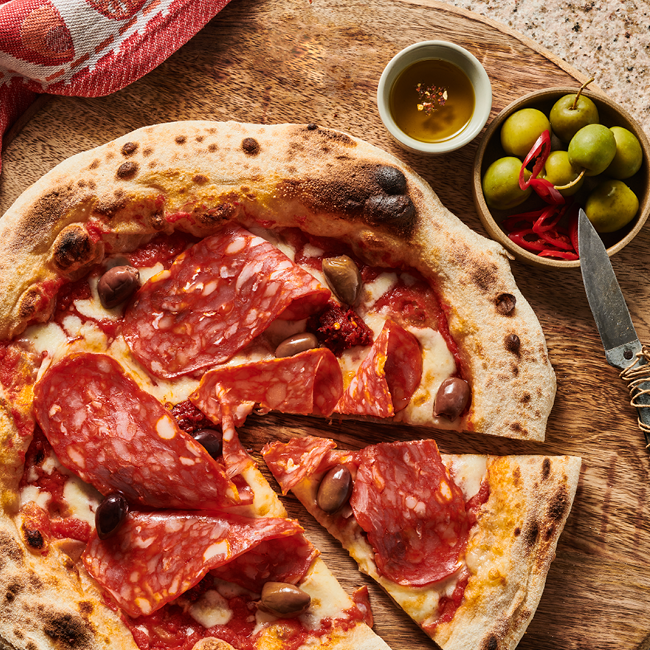 Pizza mit Salami und Oliven auf Holzbrett, daneben Oliven, Chili und ein Schälchen mit Öl, Messer und Serviette.