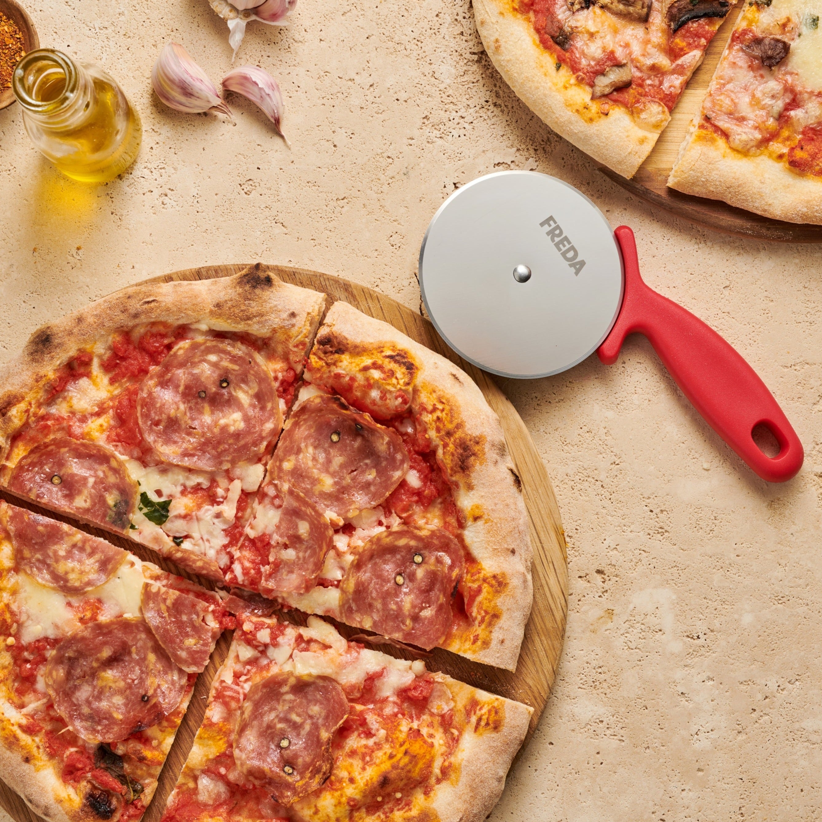 Pizza mit Salami auf Holzbrett, roter Pizzaschneider und Knoblauch auf einem beigen Hintergrund.
