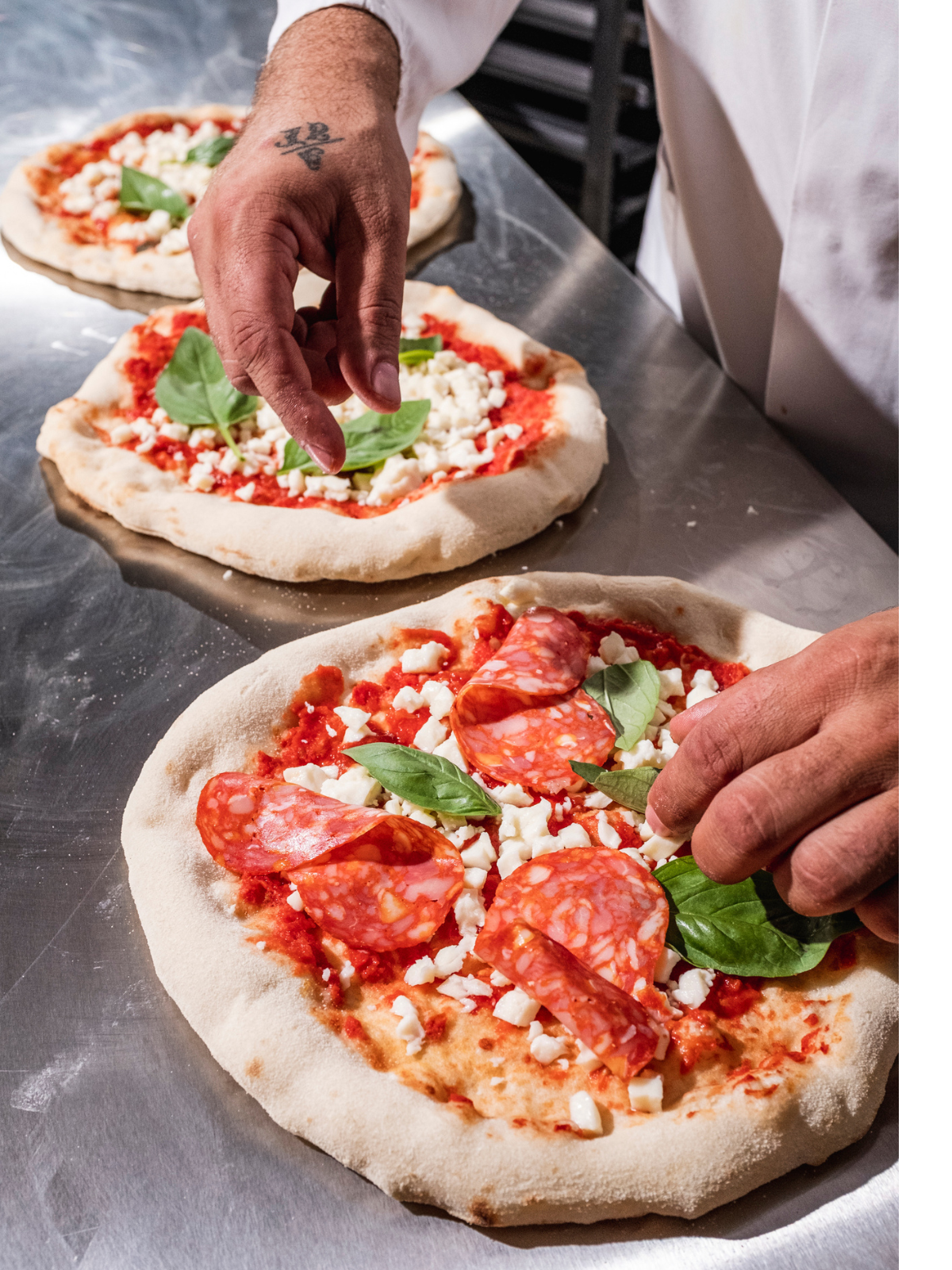 Eine Person belegt Pizzen mit Salami, Basilikum und Käse auf einer Metalloberfläche.
