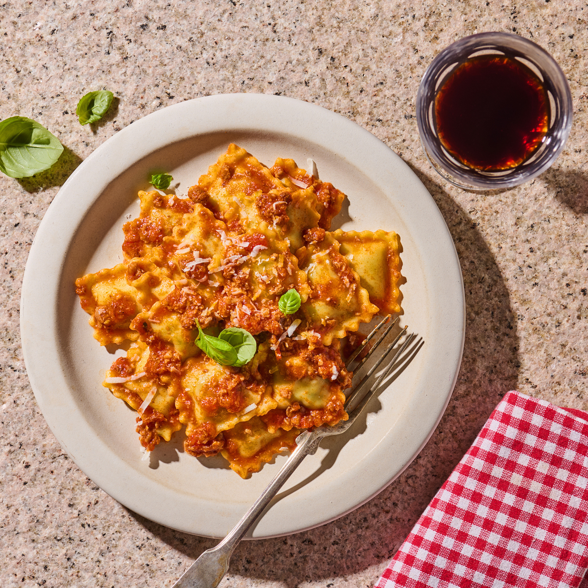 Ein Teller mit Ravioli in Tomatensauce, garniert mit Basilikum, daneben ein Glas Rotwein auf einem Tisch mit rot-weißem Tuch.