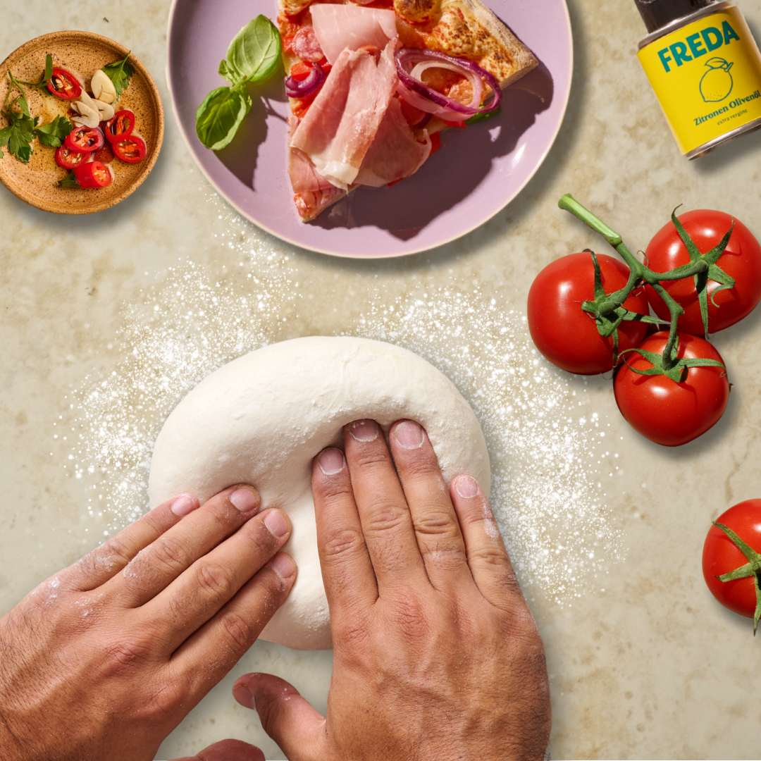 Hände kneten Teig auf bemehlter Oberfläche; daneben Tomaten, eine Pizzascheibe auf einem Teller und Gewürze.