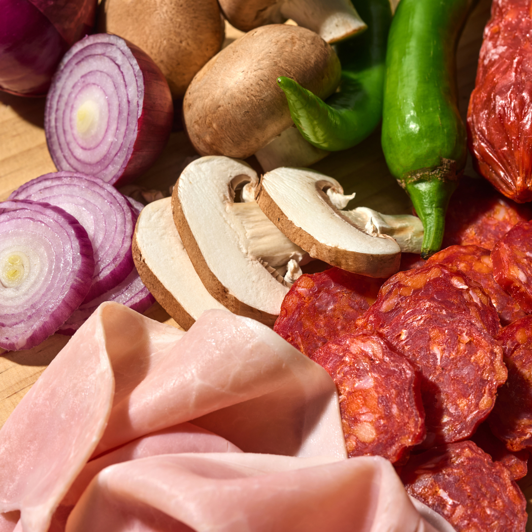 Verschiedene Pizzabeläge aus roter Zwiebel, Champignons, grünem Pfeffer, Schinken und Wurst auf einem Holztisch.