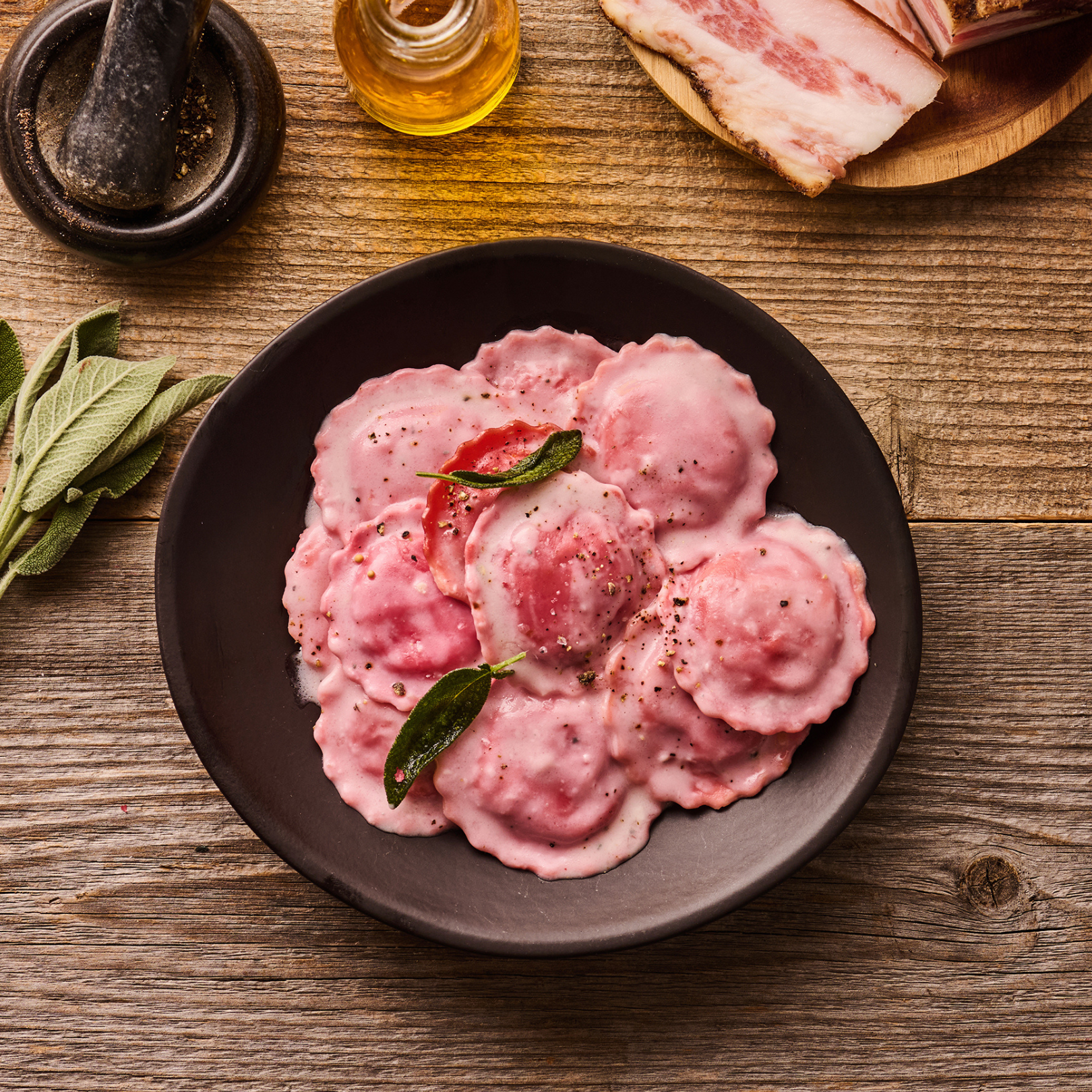 Rosa Ravioli in einer dunklen Schale, dekoriert mit Salbeiblättern, neben Olivenöl, Speck und einem Mörser auf Holztisch.
