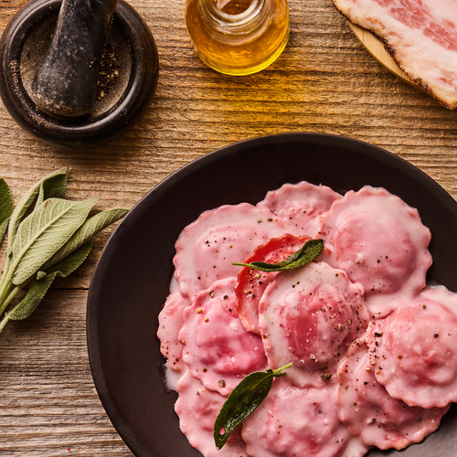 Rosa Ravioli mit Salbeiblatt, Pfeffer und Olivenöl auf einem Teller, daneben Salbeiblätter und ein Mörser.