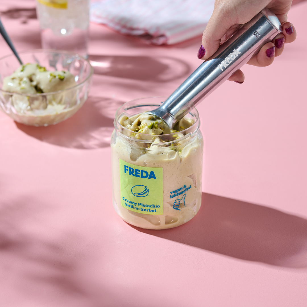 Hand holt Pistazien-Sorbet aus einem Glas mit einem Löffel auf einem rosa Tisch.