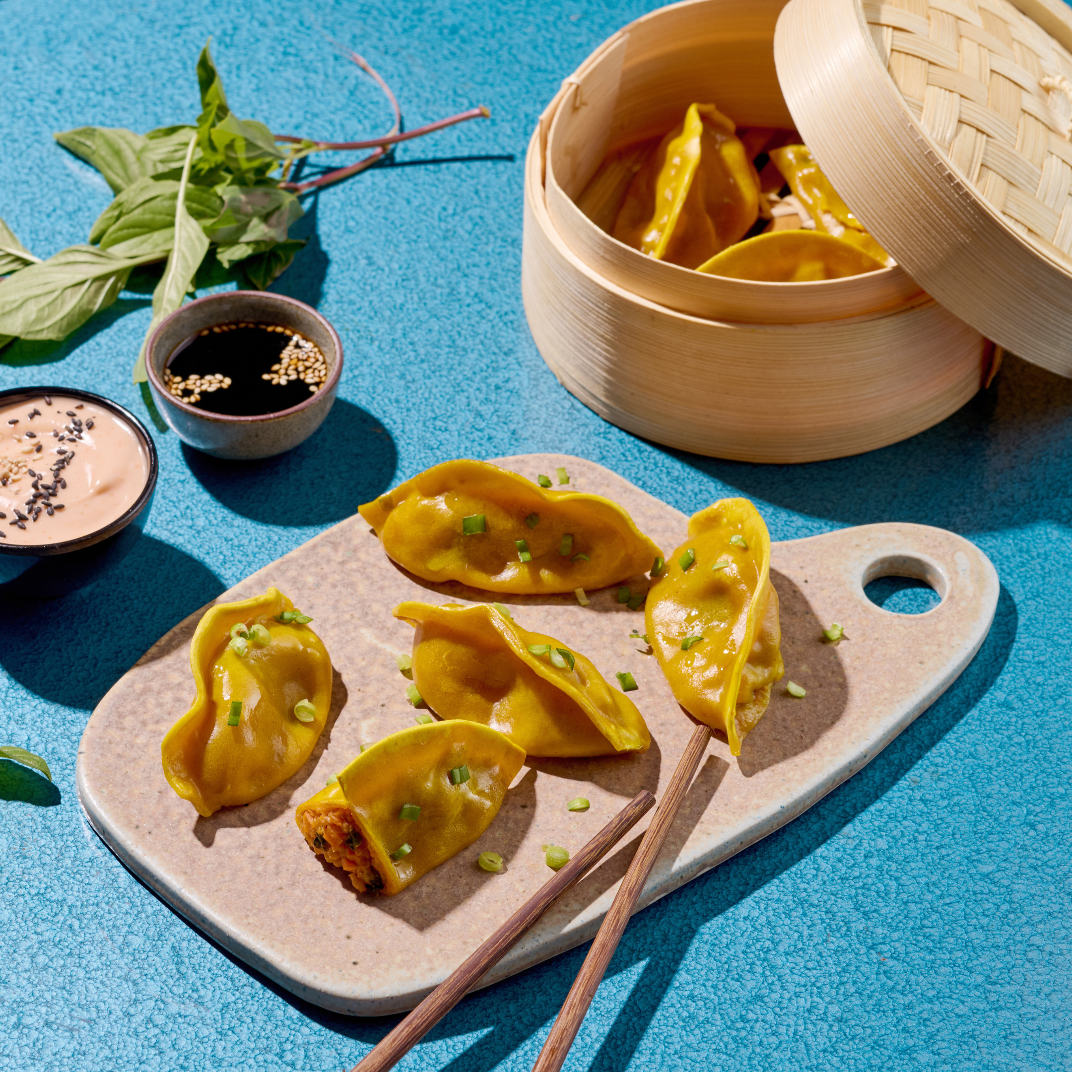 Gelbe Dumplings auf einem Steinbrett, mit Stäbchen, Sojasoße und Dip, daneben ein Bambuskörbchen auf blauem Hintergrund.