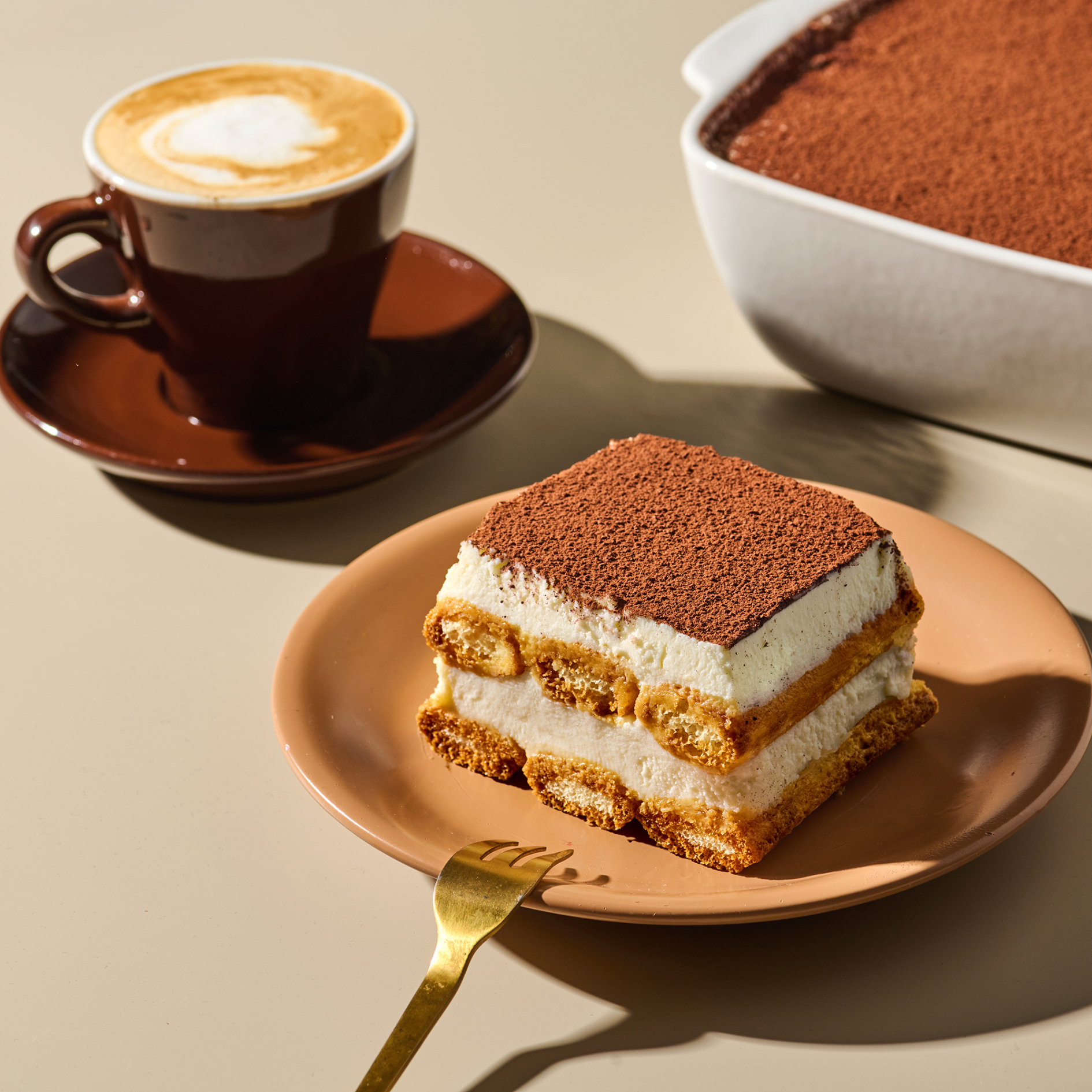 Tiramisu-Stück auf einem Teller mit einer Gabel und einer Tasse Kaffee daneben, im Hintergrund ein Tiramisu-Auflaufform.