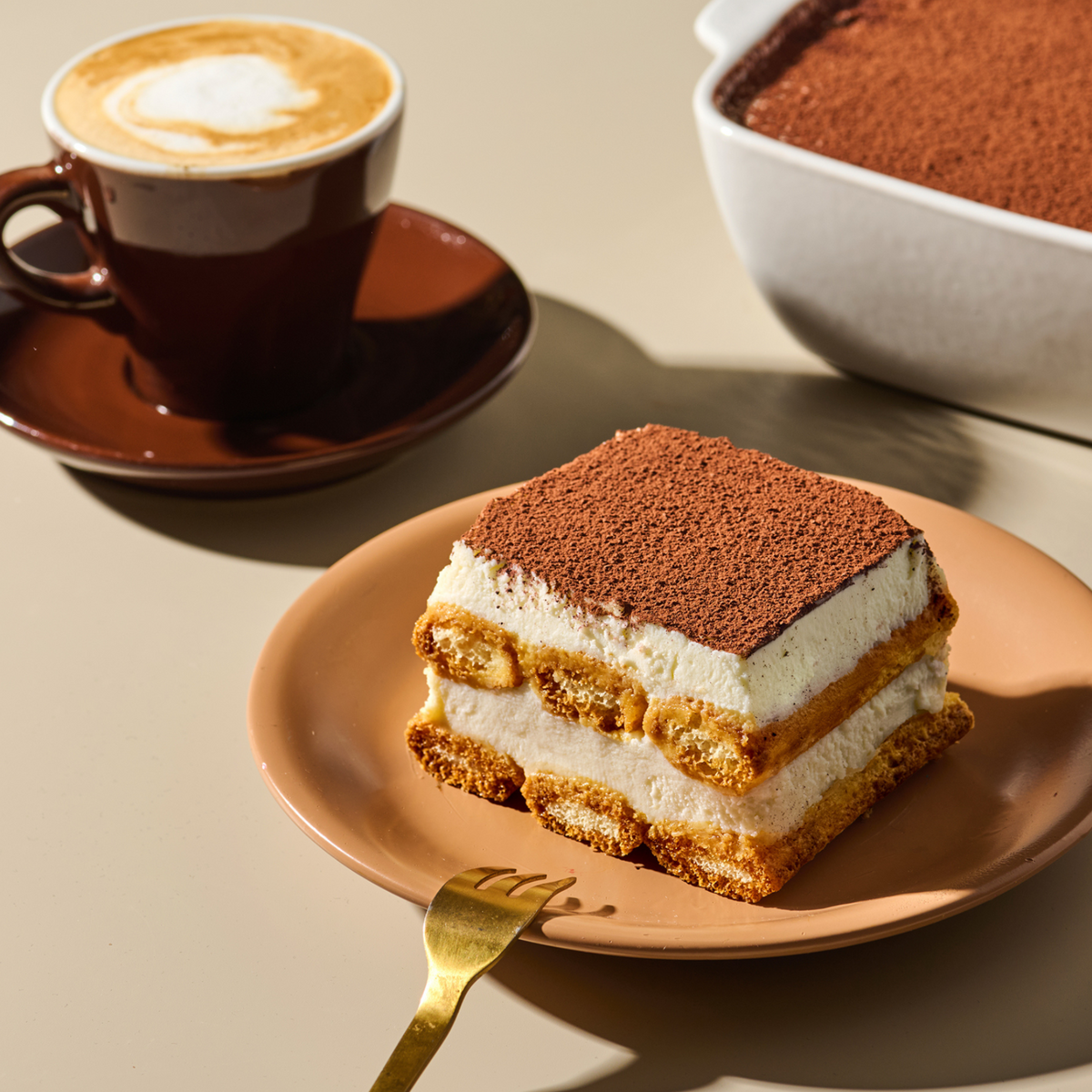 Tiramisu-Stück auf einem Teller mit einer Gabel und einer Tasse Kaffee daneben, im Hintergrund ein Tiramisu-Auflaufform.