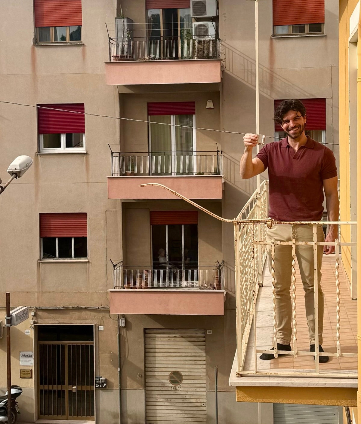 Ein Mann steht auf einem Balkon und hält ein Glas hoch, während er lächelt; im Hintergrund Wohngebäude.