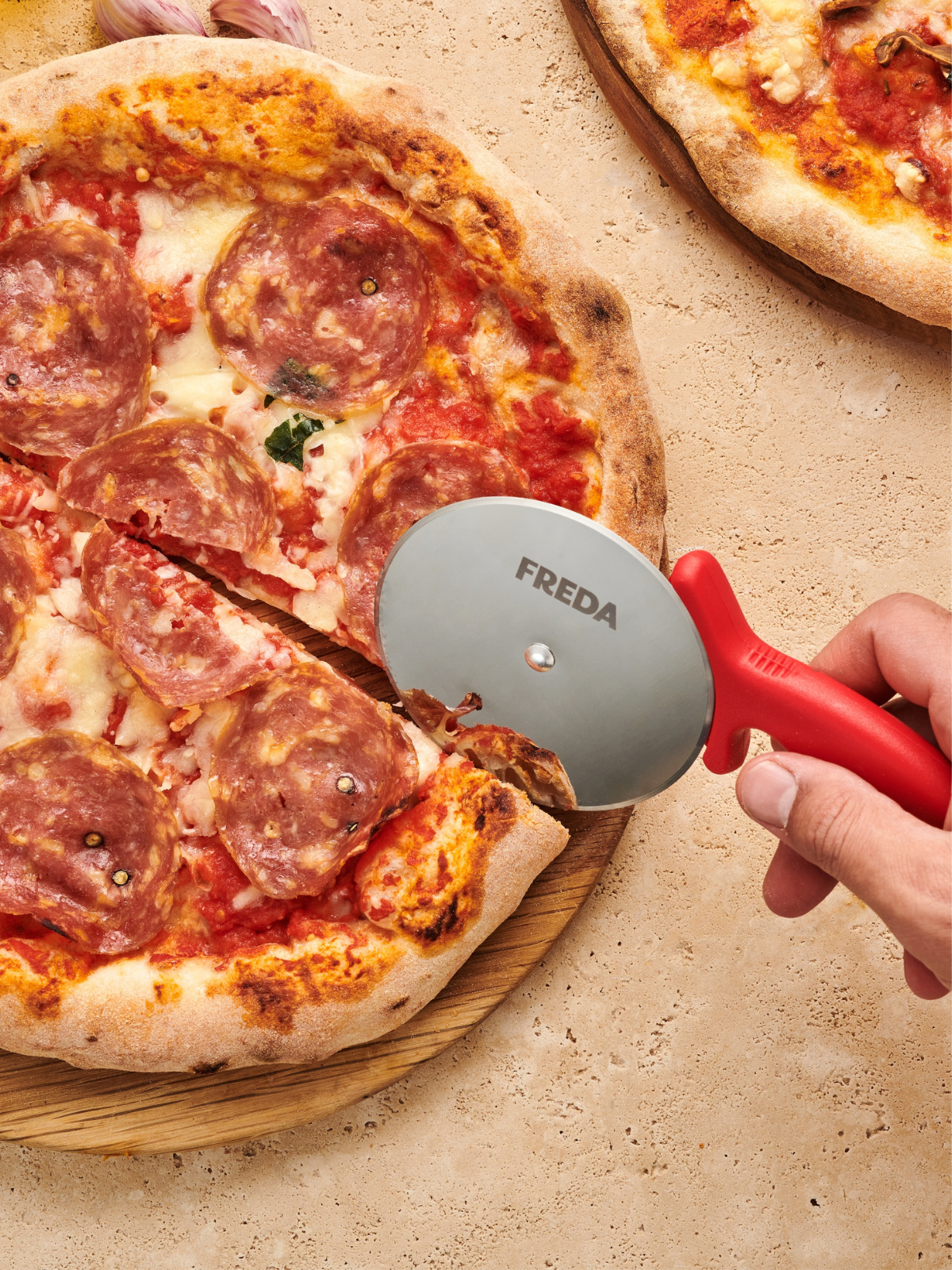 Hand schneidet Pepperoni-Pizza mit rotem Pizzaroller auf Holztisch.