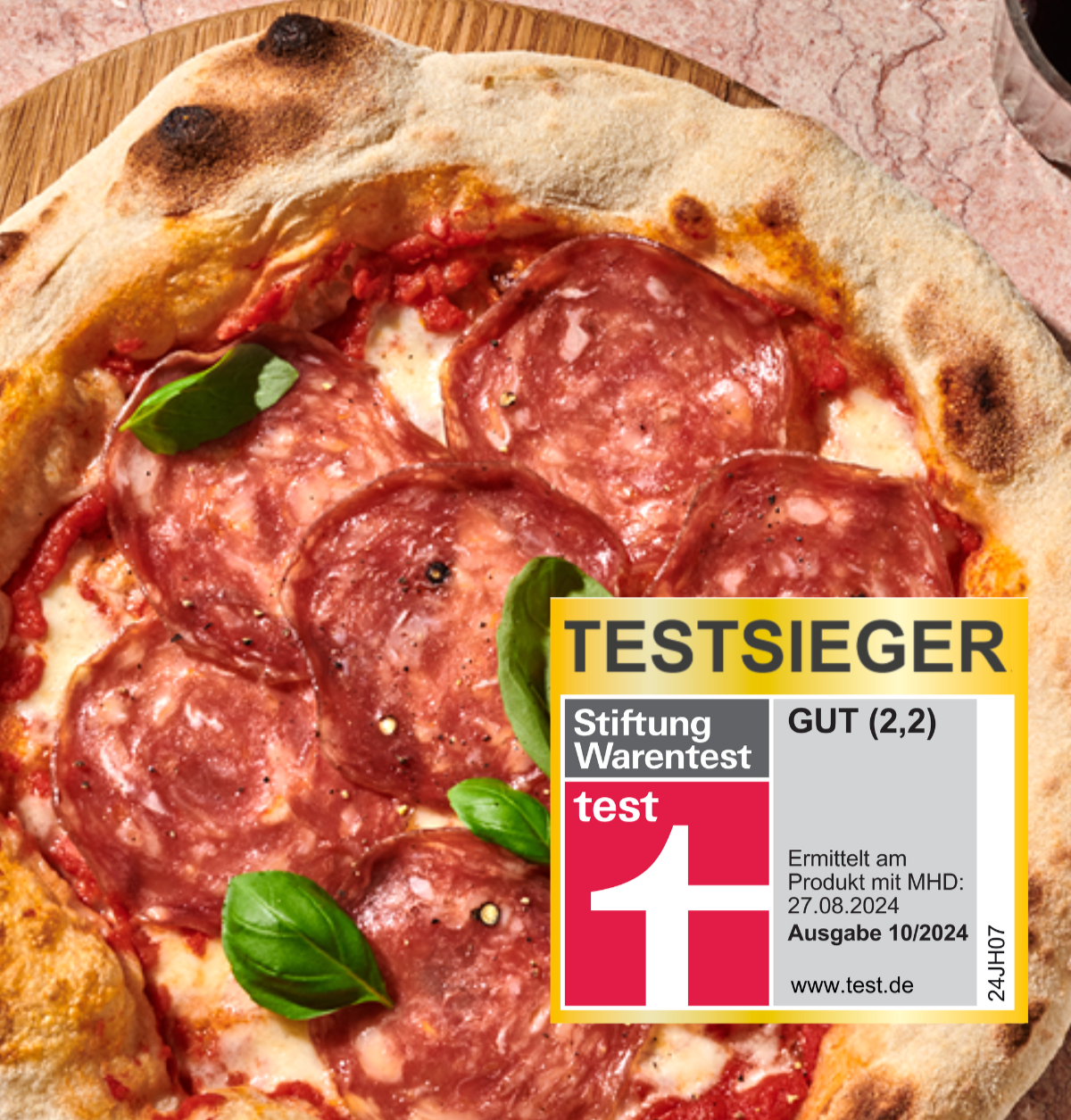 Pizza mit Salami und Basilikum auf Holzbrett, daneben Testsiegel von Stiftung Warentest mit Bewertung "GUT (2,2)".