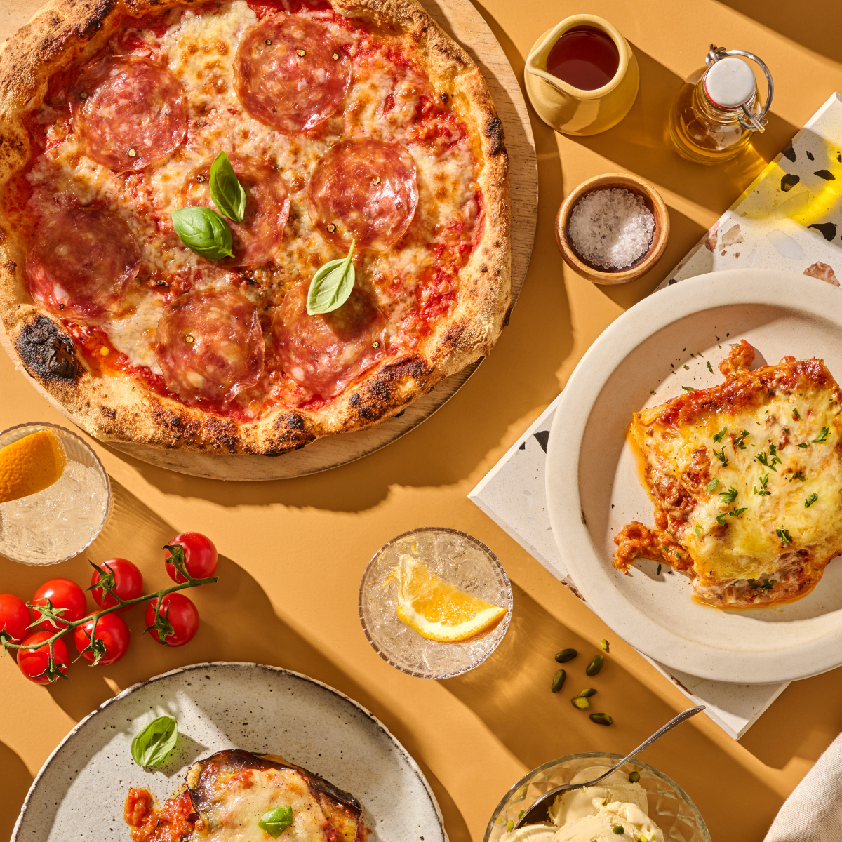 Pizza mit Salami, eine Portion Lasagne, Tomaten, Getränke und Gewürze auf einem Holztisch mit Sonnenlicht.