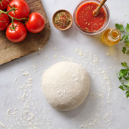 Pizzateig auf einem bemehlten Tisch mit Tomaten, Tomatensauce, Kräutern und einer Flasche Olivenöl.
