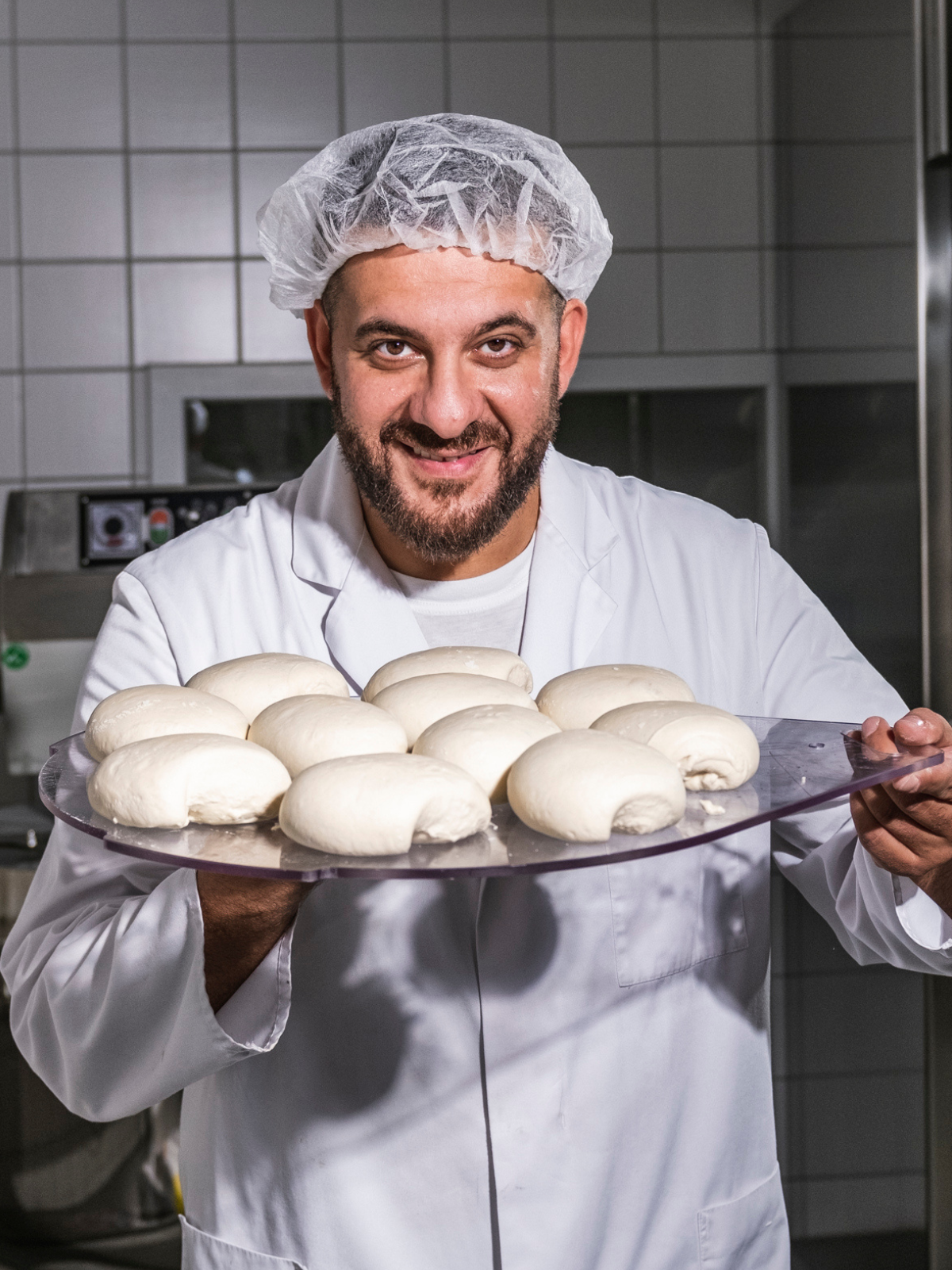 Ein Mann in weißem Kittel und Haarnetz hält ein Tablett mit Teigbrötchen in einer Küche.