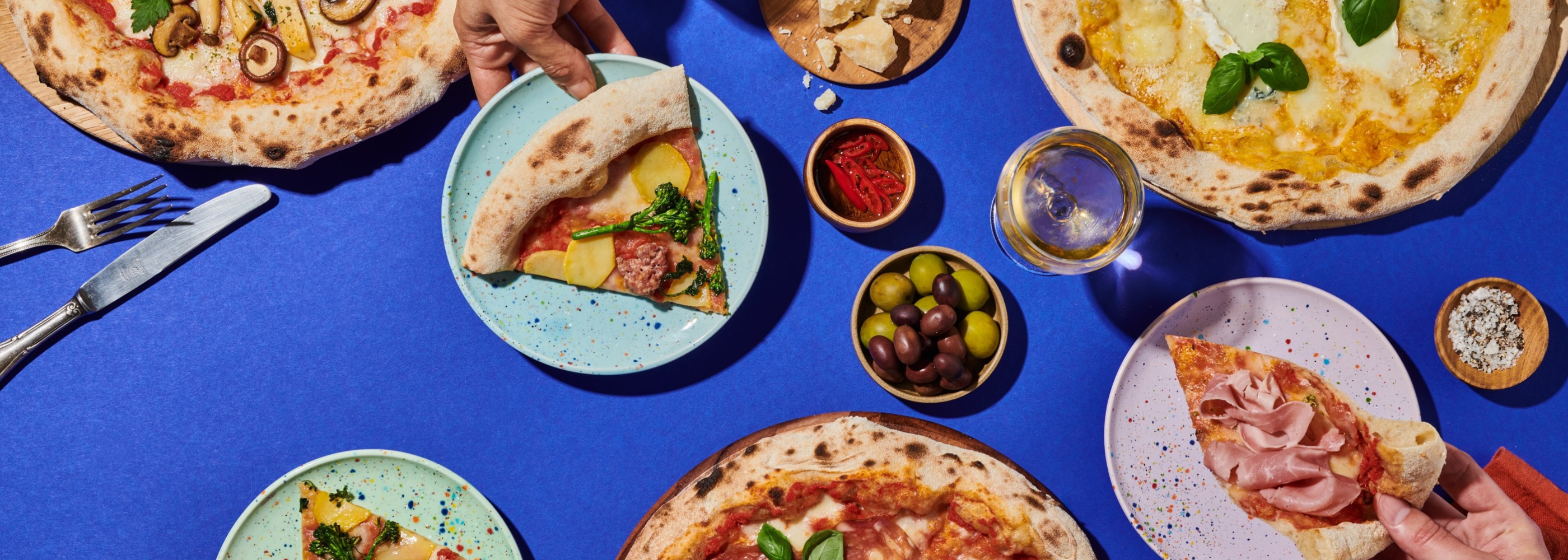 Verschiedene Pizzen auf einem blauen Tisch mit Oliven, Paprika und einem Glas Weißwein. Hände halten Pizzastücke.