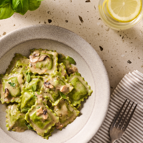 Grüne Ravioli mit Pilzsoße und Kräutern auf einem Teller. Daneben liegt eine Gabel; im Hintergrund ein Glas Wasser mit Zitrone.
