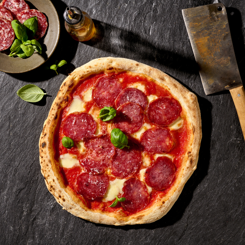 Pizza mit Salami und Basilikumblättern, daneben ein Messer und ein Teller mit Salami und Basilikum auf schwarzer Oberfläche.