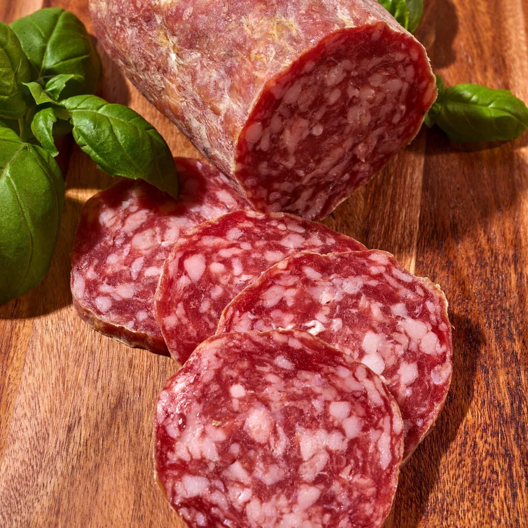 Auf einem Holztisch liegen eine halbierte Salami und einige geschnittene Scheiben, neben frischem Basilikum.