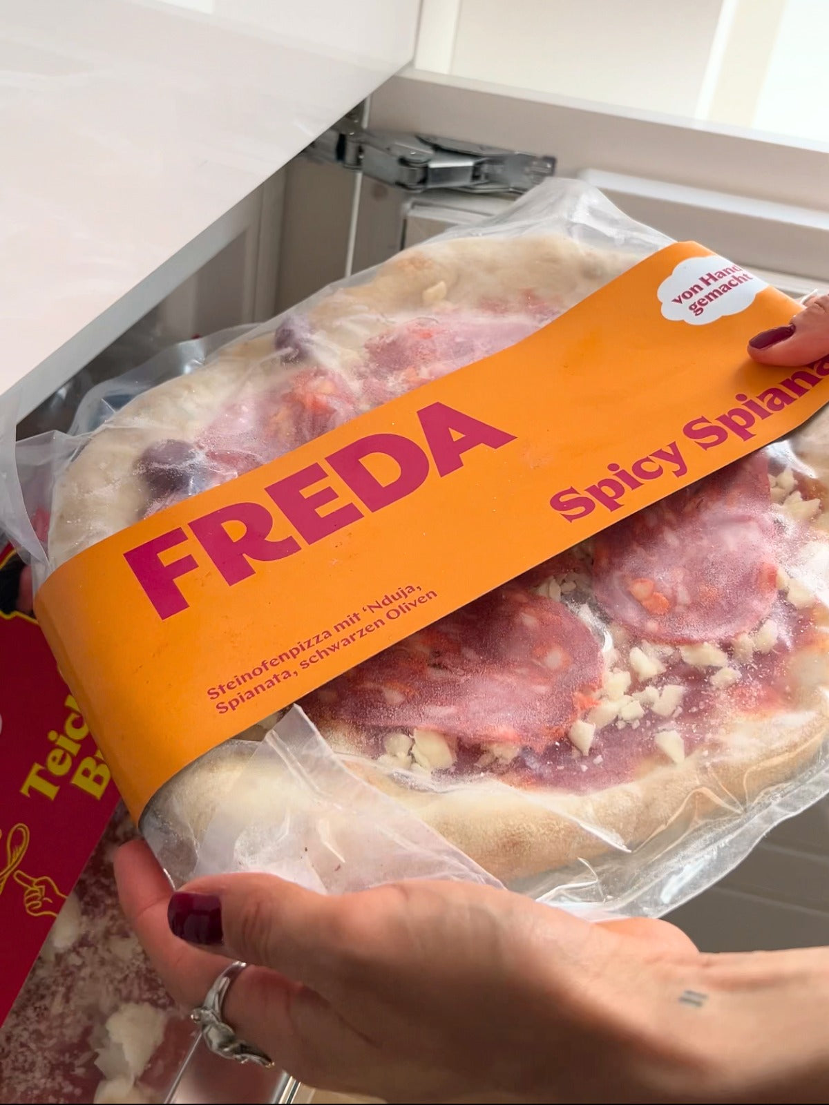 Eine Person hält eine tiefgekühlte Pizza in einer Verpackung mit der Aufschrift "FREDA Spicy Spianata".