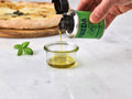 Eine Hand gießt Öl aus einer grünen Dose in ein Glas, daneben liegt Basilikum, im Hintergrund eine Pizza.