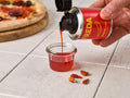 Hand gießt Chili-Olivenöl aus einer Dose in ein Glas, mit Pizza und Chilischoten im Hintergrund.