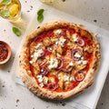 Pizza mit Hähnchen, roten Zwiebeln und Feta auf einem weißen Untergrund, daneben Pfefferflocken, ein Zitronenstück und getränke.