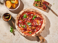 Pizza mit Paprika und Basilikum auf einem Holzbrett, daneben ein Getränk mit Zitrusfrüchten und ein Holzbrett mit Oliven.