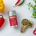 Eine Dose "FREDA Paprika Chili" liegt auf einem Tisch, umgeben von Paprika, Chilischoten und Petersilie.