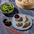 Dampfnudeln und Edamame auf Marmortisch, serviert mit Sojasauce und Chili-Dip, rote Essstäbchen daneben.