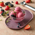 Eine Schüssel mit Beeren-Eiscreme, garniert mit Brombeeren und Thymian, umgeben von frischen Beeren und einem Eislöffel.