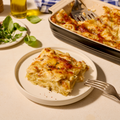 Ein Stück Lasagne auf einem Teller neben einer Auflaufform mit mehr Lasagne und frischen Basilikumblättern.