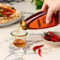 Eine Hand gießt Honig aus einer Flasche in ein Glas vor einem Teller mit roten Chilischoten und einer Pizza.