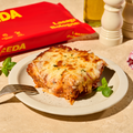 Ein Teller mit einem Stück Lasagne Bolognese, daneben eine Packung Lasagneblätter und eine Pfeffermühle.
