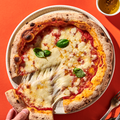 Pizza mit geschmolzenem Käse und Basilikumblättern, eine Hand nimmt ein Stück, daneben eine Tasse Olivenöl.