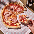 Eine Pizza mit Schinken auf kariertem Papier, eine Hand nimmt ein Stück auf, daneben steht eine Flasche.