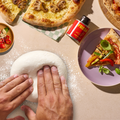 Hände kneten Teig mit Pizzascheiben, Chiliöl und Gewürzen auf einem Tisch.