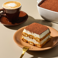 Tiramisu-Stück auf einem Teller mit einer Gabel und einer Tasse Kaffee daneben, im Hintergrund ein Tiramisu-Auflaufform.