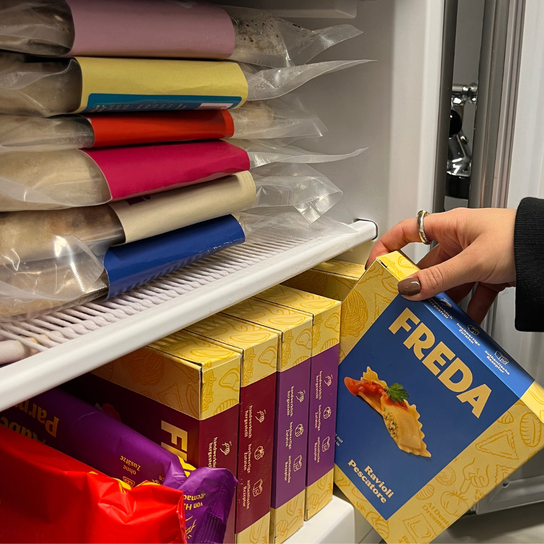 Hand greift nach einer blauen Box mit Essen aus einem gut gefüllten Gefrierschrank.